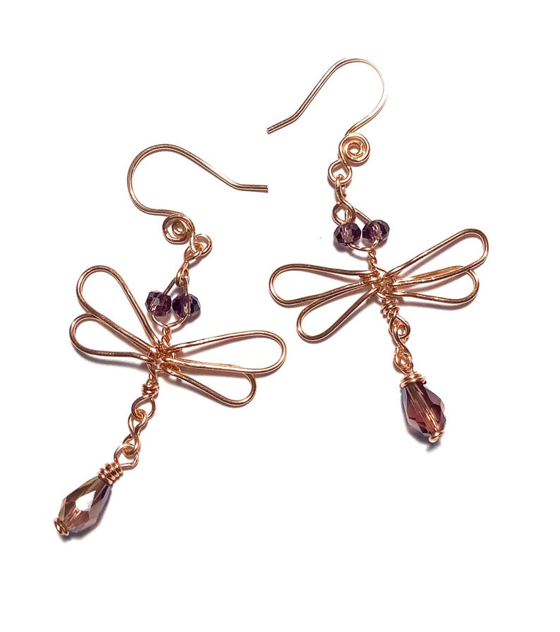 May include: A pair of copper-colored dragonfly earrings. Each earring features a dragonfly design with wire wings, small purple beads, and a dangling faceted purple crystal. The earrings have a hook closure.