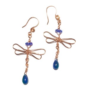 May include: A pair of copper-colored wire dragonfly earrings. Each earring features a dragonfly design with blue bead accents and a teardrop-shaped blue bead dangling below. The earrings have a hook closure.