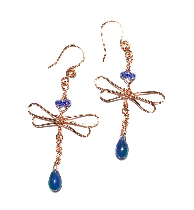 May include: A pair of copper-colored wire dragonfly earrings. Each earring features a dragonfly design with blue bead accents and a teardrop-shaped blue bead dangling below. The earrings have a hook closure.