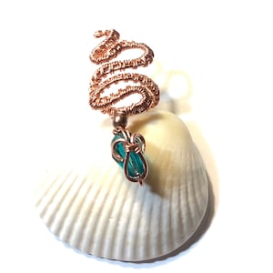 May include: A copper wire pendant with a spiral design and a teal crystal bead. The pendant is displayed on a white seashell. The handcrafted wirework and the teal crystal create a striking contrast against the shell.