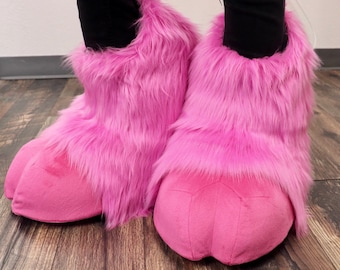 May include: A pair of pink fuzzy slipper boots with a claw-like toe. The boots are made of soft, plush fabric and have a pink fur trim.