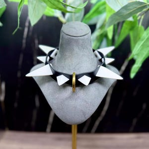 Puede incluir: Un collar de púas en blanco y negro con un anillo dorado, exhibido en una cabeza de maniquí gris. El collar presenta formas geométricas blancas y negras alternas y púas blancas. El fondo incluye follaje verde.