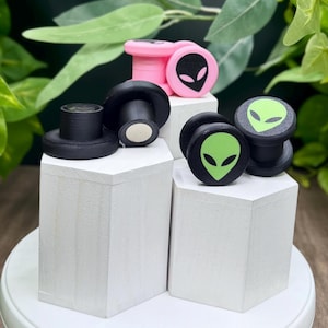 May include: A collection of ear plugs in various colors, including black and pink. Each plug features a green or black alien head design. The plugs are displayed on white geometric blocks against a backdrop of green foliage.