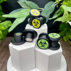 Puede incluir: Taponas para los oídos negras con símbolos de peligro biológico amarillos y verdes. Las taponas se exhiben sobre pedestales geométricos blancos, rodeadas de follaje verde. Las taponas son de un material oscuro.