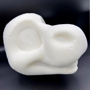 May include: A white, abstract sculpture with rounded forms and smooth surfaces. The object has a unique shape, possibly resembling a stylized head or a natural form. The sculpture is set against a dark background.