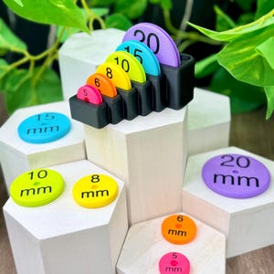 May include: A black plastic organiser stand holding a set of colourful circular gauges in sizes 5, 6, 8, 10, 15, and 20 mm. Additional matching circular gauges are displayed on white hexagonal blocks, each clearly labelled with its respective size in mm.