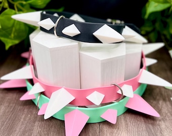Può includere: Tre collari regolabili in nero, rosa e verde menta, ciascuno caratterizzato da una serie di punte bianche e da un anello a D in metallo argentato. I collari sono impilati verticalmente, mostrando una struttura a fascia liscia e flessibile.