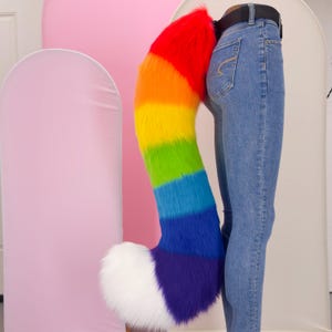 Pode incluir: Uma longa cauda de pelo sintético felpudo com um gradiente de arco-íris vibrante em vermelho, laranja, amarelo, verde, azul e roxo, terminando numa ponta branca sólida. A cauda está presa a um cinto preto sobre calças de ganga azul-claras.