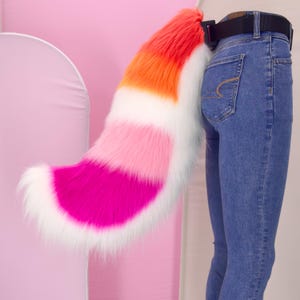 May include: A colorful faux fur tail in shades of red, orange, white, pink, and magenta. The tail is attached to a black belt worn with blue jeans. The tail is fluffy and has a curved shape.
