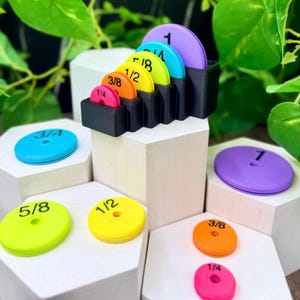 Puede incluir: Un conjunto de discos de medición circulares de plástico en varios colores, incluyendo morado, azul, verde lima, amarillo, naranja y rosa. Cada disco tiene marcada una fracción o número entero en negro: 1/4, 3/8, 1/2, 5/8, 3/4 y 1. Un soporte negro los organiza.