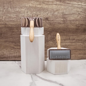 May include: Two wooden handled grooming brushes with metal bases. The brush on the left has a copper colored base and the brush on the right has a gray bristle brush head.