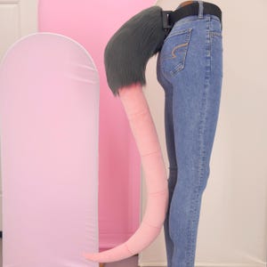 May include: A long, segmented, light pink tail accessory attached to a black belt. The base of the tail features thick, dark gray faux fur. The tail is displayed on a pair of blue denim jeans, showing a smooth, cylindrical, and flexible design.