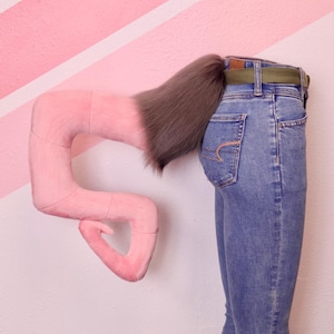 May include: A pink, plush, curved object with a brown, furry extension, positioned near the back of a person wearing blue jeans with a green belt. The background features a pink and white striped wall.