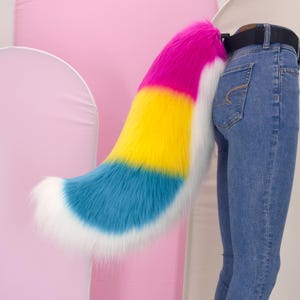 Pode incluir: Uma cauda colorida de pele sintética em um gradiente de rosa, amarelo, azul e branco. A cauda é presa a um cinto preto usado com jeans azuis. A cauda é um acessório de fantasia.