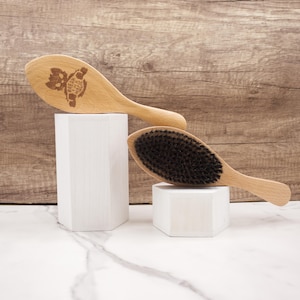 May include: Two wooden hair brushes on white pedestals. The brush on the left has a wooden handle and a rounded head with the text "Freak The Fade" engraved on it. The brush on the right has a black bristle head and a wooden handle.
