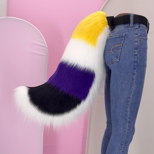 Puede incluir: Una cola de felpa con los colores de la bandera del orgullo no binario, amarillo, blanco, morado y negro. La cola está sujeta a un cinturón negro que se lleva con vaqueros azules. La cola es esponjosa y tiene un degradado de color.