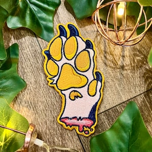 May include: Embroidered patch featuring a stylized paw print with yellow pads, blue claws, and a pink and red blood-like detail. The patch is outlined in a cream color. Decorative leaves and string lights surround the patch.
