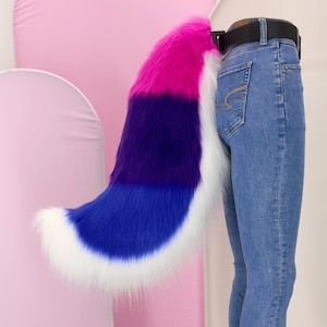 Puede incluir: Una cola de piel sintética con un diseño de bloques de color. La cola presenta un degradado de rosa, morado, azul y blanco. La cola está sujeta a un cinturón negro. Accesorio de disfraz.