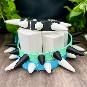 Può includere: Tre collari impilati caratterizzati da una fascia liscia e grandi punte acuminate. Il collare superiore è nero con punte bianche, quello centrale è verde menta con punte nere e quello inferiore è blu brillante con punte bianche. Ognuno ha un anello a D.