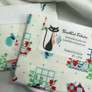 May include: Two folded fabric fat quarters with a white background and a colorful pattern of houses, flowers, and chickens. One quarter has a label that reads "BratKat Fabrics" and "Standard Fat Quarter 18" x 22""