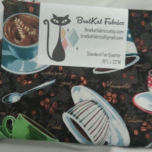 May include: A fabric square with a coffee-themed print featuring coffee cups, beans, and text. The fabric is dark with a white label that includes the text "BratKat Fabrics" and the dimensions of the fabric: 18"L x 22"W.