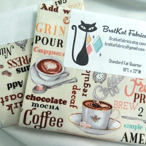 May include: Fabric bundles with a coffee theme. The fabric features coffee cups, coffee beans, and text like "French Press" and "Cappuccino." A business card with a black cat logo is on top. The fabric measures 18" x 22".