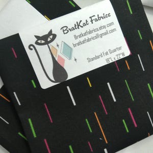 May include: Black fabric with a colorful vertical stripe pattern. A white label features a black cat logo and the text "BratKat Fabrics". The fabric is a Standard Fat Quarter, measuring 18" x 22".
