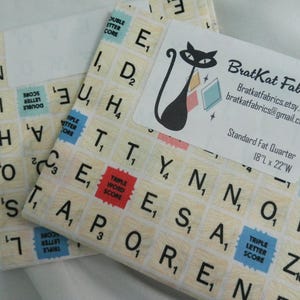 May include: Two fabric bundles with a Scrabble tile pattern. The fabric is cream-colored with black letters and numbers, and red, blue, and pink score squares. One bundle has a label with a black cat logo and the text "BratKat Fabrics".