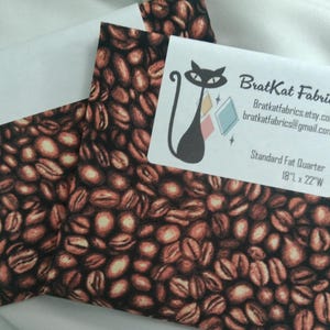 May include: Two fabric pieces with a coffee bean pattern. The fabric is brown and tan with a repeating design of coffee beans. A white label with a black cat logo and text is on top of the fabric. The label reads "BratKat Fabrics" and includes contact information.