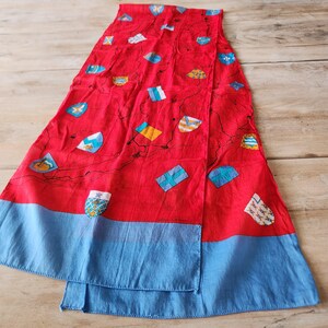 May include: A rectangular scarf featuring a vibrant red background with a scattered pattern of various colorful heraldic shields and crests. The scarf is finished with a solid light blue border along the bottom edge and thin black lines.