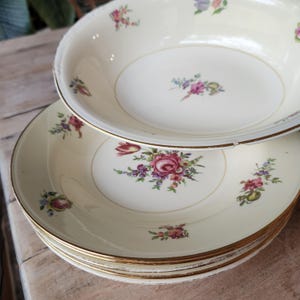 May include: A stack of cream-colored vintage plates and a bowl with gold rims and floral designs. The plates and bowl feature a central floral arrangement with pink and purple roses, and smaller floral accents.