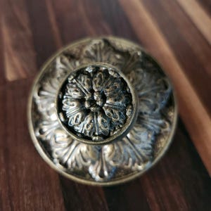 May include: Close-up of an antique brass cabinet knob. The knob features a central floral design surrounded by ornate, embossed detailing. The finish has a weathered, aged appearance, with darker accents in the recessed areas, giving it a vintage aesthetic.