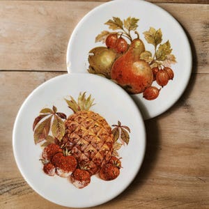 May include: Two white ceramic plates with fruit illustrations. One plate features a pineapple and strawberries, the other a pear and berries. The plates have a vintage aesthetic with detailed, colorful fruit designs.