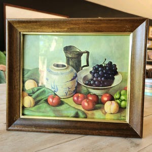 May include: Framed still life art print featuring a composition of fruit and pottery. The artwork includes a bowl of grapes, tomatoes, peaches, and a decorative vase and pitcher. The frame is a dark brown wood with a gold inner border.