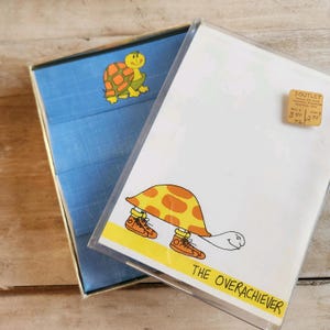May include: A vintage stationery set featuring a cartoon turtle with a yellow and orange shell wearing orange sneakers. The text "THE OVERACHIEVER" is below the turtle. The set includes a blue fabric-covered box with a turtle graphic.