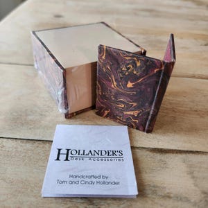 May include: A set of desk accessories with a marbled design in shades of burgundy and gold. The set includes a box and a small, open container. A white label reads "Hollander's Desk Accessories" and "Handcrafted by Tom and Cindy Hollander."