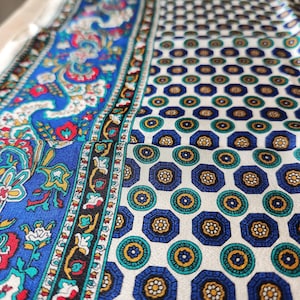 May include: A fabric scarf featuring a split design with a vibrant blue section displaying a multi-colored paisley pattern and a cream-colored section decorated with a repeating geometric motif of blue octagons and small yellow floral centers.