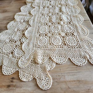 May include: An off-white crochet table runner featuring a repeating floral motif pattern. The textile has a scalloped edge border with intricate openwork stitching throughout the rectangular design, showcasing a detailed needlework construction.