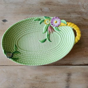 May include: A light green oval serving platter with a woven basket design. The platter features a yellow handle and is decorated with colorful flowers and green leaves. The flowers include purple, yellow, and pink blossoms.