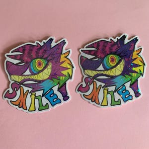 May include: Two stickers featuring a cartoon cat with a wide grin and rainbow-colored eyes. The cat's fur is purple, pink, and yellow. The word "SMILE" is written in rainbow colors below the cat's face. The stickers have a holographic effect.