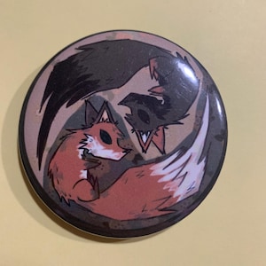 May include: A round button with a cartoon fox and raven design. The fox is orange and white, while the raven is black. The background is a mix of brown and tan. The button has a glossy finish.