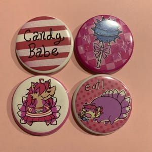 May include: Four circular badges with cartoon designs. One has red and white stripes and the words "Candy Babe." Another features a lollipop with a bow. The other two badges have cartoon characters and the word "eat!"