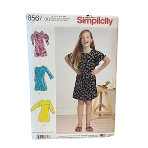 May include: Simplicity sewing pattern 8567 for girls' plus sizes 8 1/2-16 1/2. The envelope displays four dress variations: a floral cold-shoulder dress, a blue long-sleeve dress, a yellow long-sleeve dress, and a black floral short-sleeve dress.