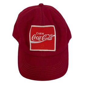 May include: A red baseball cap with a white embroidered patch that says "Enjoy Coca-Cola" in a white font.