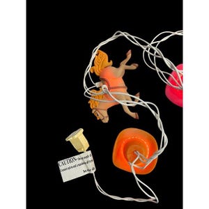 May include: String lights with carousel horse, cowboy hat, and heart-shaped decorations. The horse is orange and tan, the hat is orange, and the hearts are pink. A small tag reads "CAUTION-Do not overload".