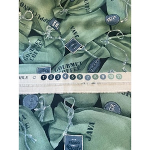 May include: Fabric with a repeating pattern of green coffee bean sacks. Each sack is labeled with coffee-related text such as "JAVA", "GOURMET COFFEE", and "CAPPUCCINO". A ruler with inch markings is visible.