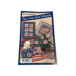 Op de afbeelding: Boekomslag "Freedom The Little Quilt Collection" met verschillende quiltontwerpen met Amerikaanse vlagmotieven. De ontwerpen omvatten kleine quilts, kussens en een miniatuurboom versierd met vlaggen. Een teddybeer in een matrozenpak is ook aanwezig.