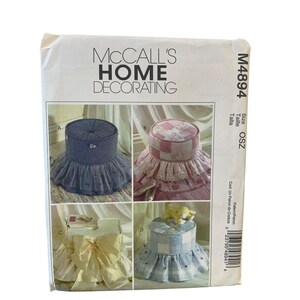 May include: A McCall's Home Decorating sewing pattern, M4894, featuring four different home decorating projects. The projects include a blue denim ottoman with a tulle skirt, a pink patchwork ottoman, a table with a yellow bow, and a blue plaid ottoman.