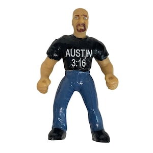 May include: A plastic action figure of a wrestler wearing a black shirt with "Austin 3:16" printed on it, blue jeans, and black shoes. The figure has a shaved head, a beard, and a serious expression.