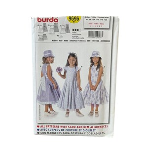 May include: A Burda 9696 sewing pattern envelope featuring illustrations of three different dress styles and a hat. The front displays three children wearing these garments in shades of light purple and white, with text including sizes 92 to 122.